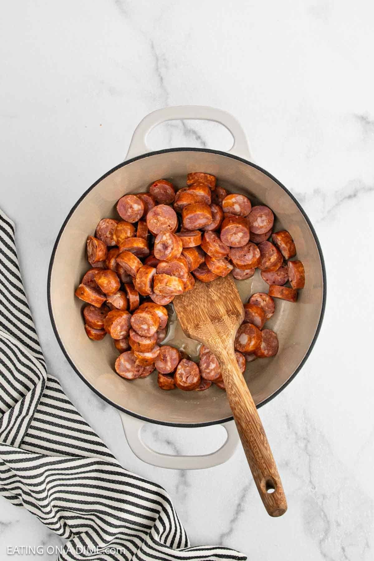 A white pot filled with sliced sausage pieces and a wooden spoon, perfect for starting your Chicken Gumbo Recipe, sits on a marble countertop next to a black and white striped kitchen towel.