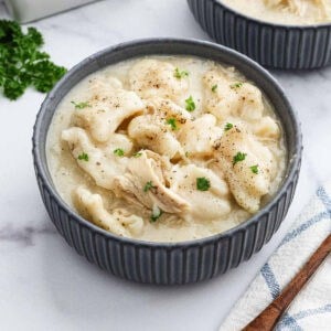 A gray bowl filled with creamy chicken and dumplings, reminiscent of classic Chicken and Biscuits, is garnished with chopped parsley and cracked black pepper. It sits on a white surface beside a striped cloth and a sprig of parsley.