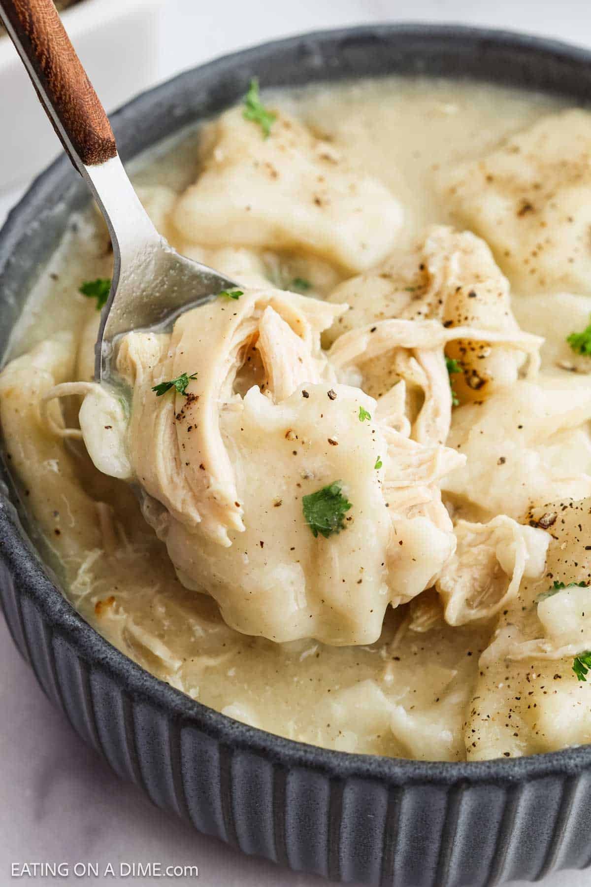 A bowl of creamy chicken and dumplings with pepper and parsley, featuring a spoon lifting tender shredded chicken and biscuit-like dumpling pieces.