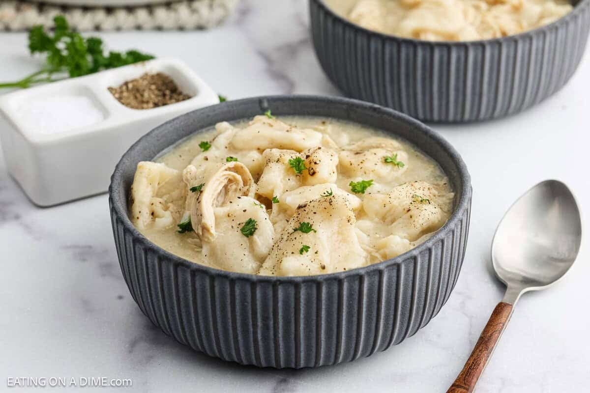 A gray bowl filled with creamy Chicken and Dumplings with Biscuits, topped with chopped parsley and black pepper, sits on a white surface next to a spoon and a small dish of salt and pepper. Another bowl is in the background.