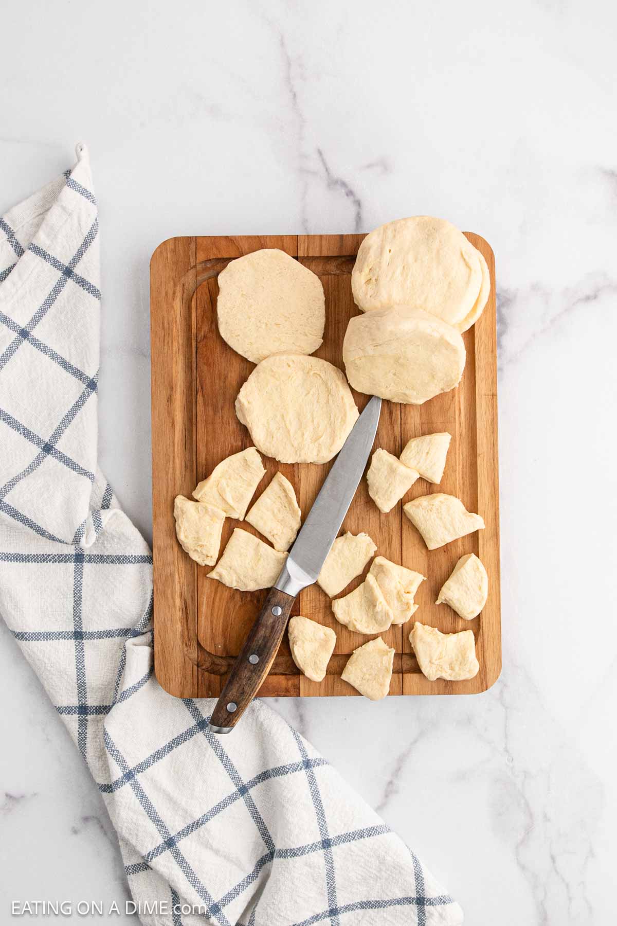 A wooden cutting board with whole and cut biscuit dough pieces, perfect for making Chicken and Dumplings with Biscuits, a knife, and a white kitchen towel with blue stripes on a marble surface.