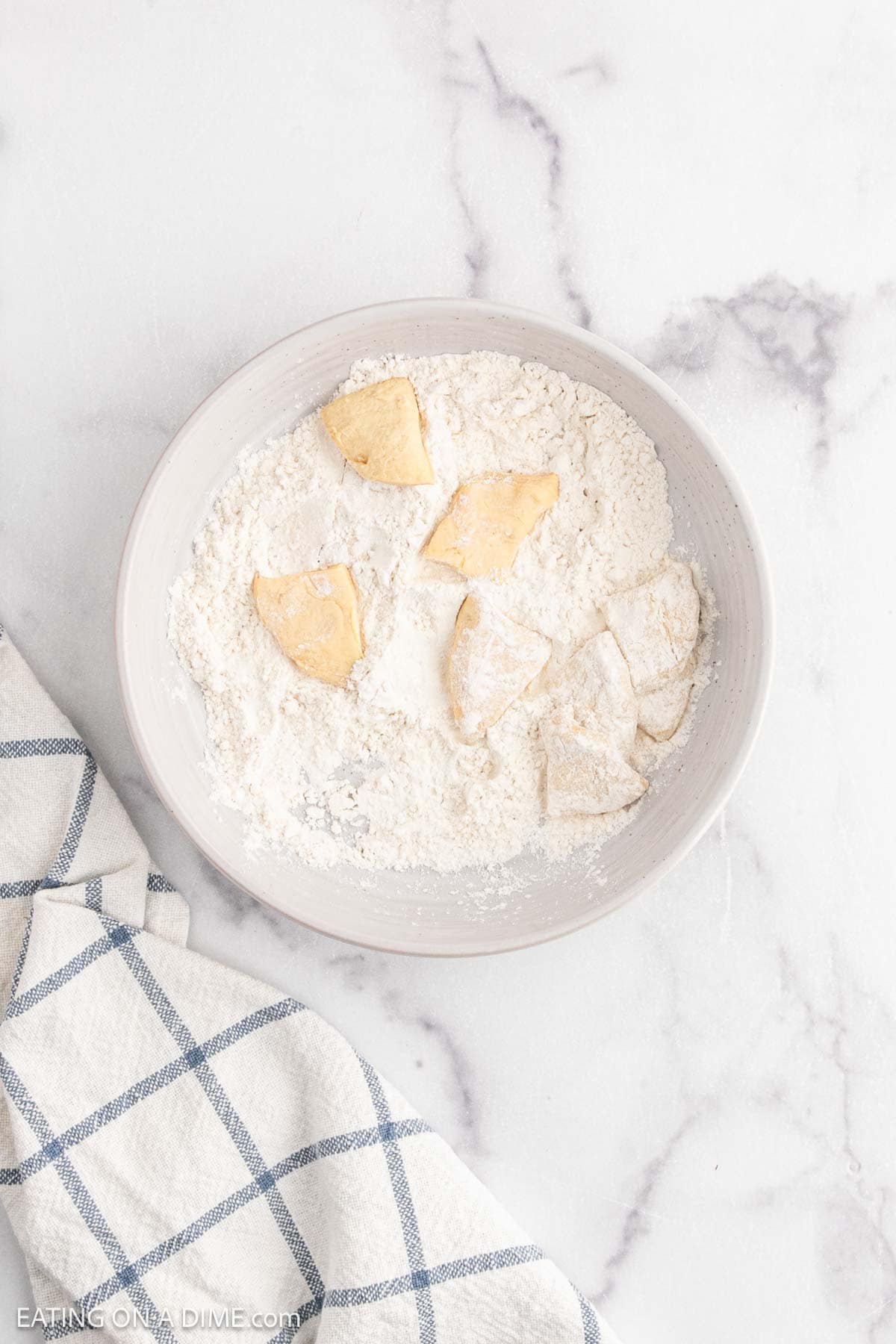 A white bowl containing dough pieces coated in flour, perfect for making Chicken and Dumplings, rests on a marble surface next to a blue and white checkered cloth.