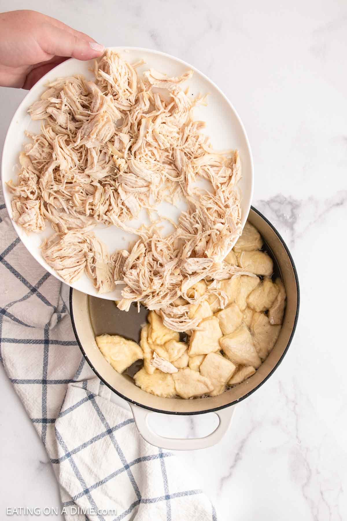 Shredded chicken is being added from a white plate into a pot containing biscuit dough pieces, creating a delicious Chicken and Biscuits dish, set on a white surface with a blue checkered towel beside it.