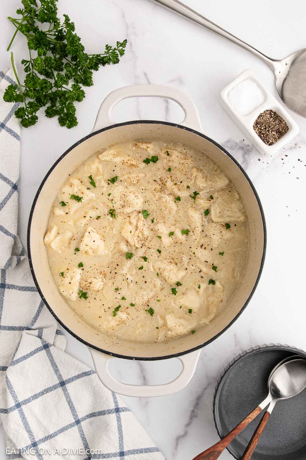 A pot of creamy chicken and dumplings garnished with chopped parsley, surrounded by a napkin, fresh parsley, salt and pepper, a ladle, and small plates on a marble countertop—perfect comfort food like classic chicken and biscuits.