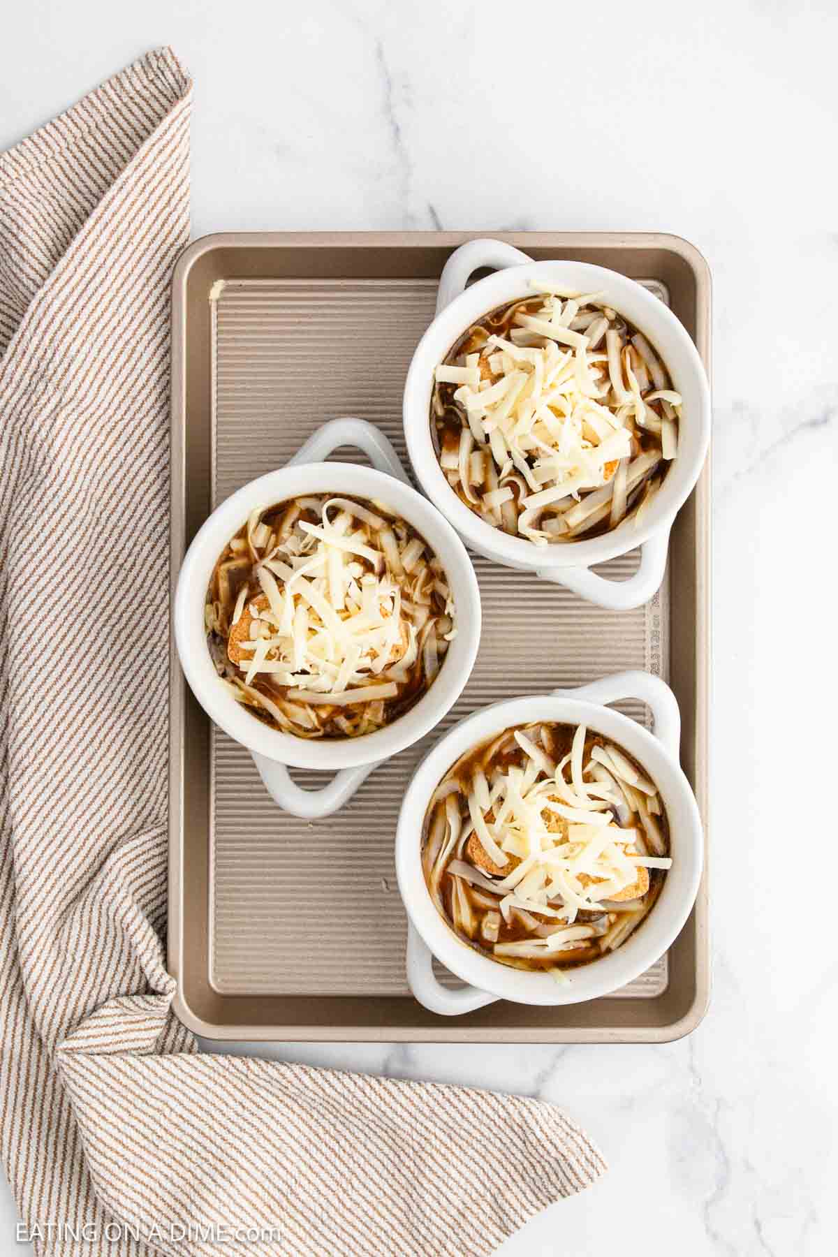 Three white ramekins filled with French Onion Soup, topped with shredded cheese, are arranged on a baking sheet next to a striped cloth napkin on a marble surface.