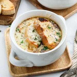 A white bowl of French Onion Soup topped with melted cheese and two toasted baguette slices, garnished with chopped parsley, sits on a wooden plate.