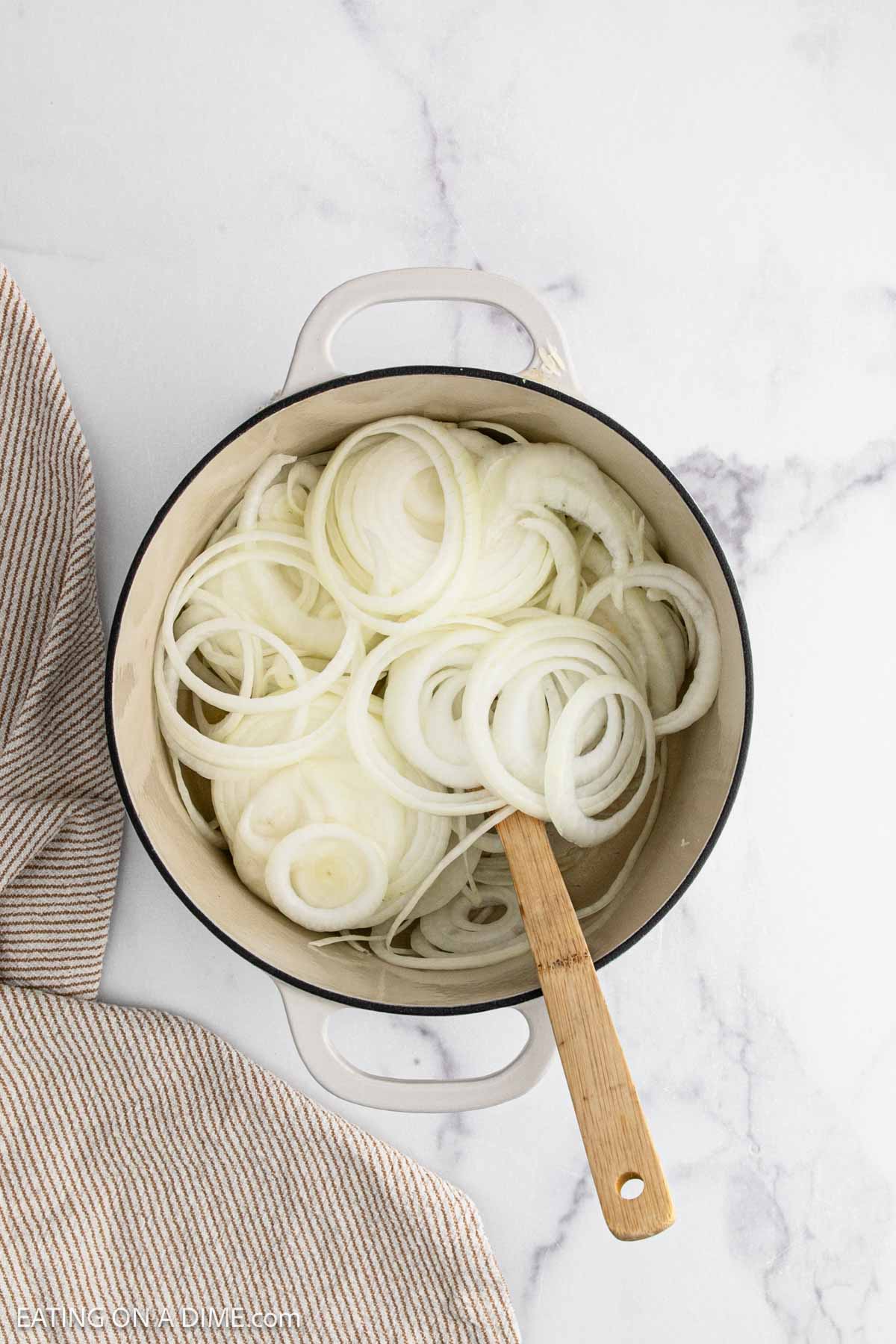 A white pot filled with raw, sliced onions and a wooden spoon, ready to start French Onion Soup, sits on a white marble surface next to a striped cloth.