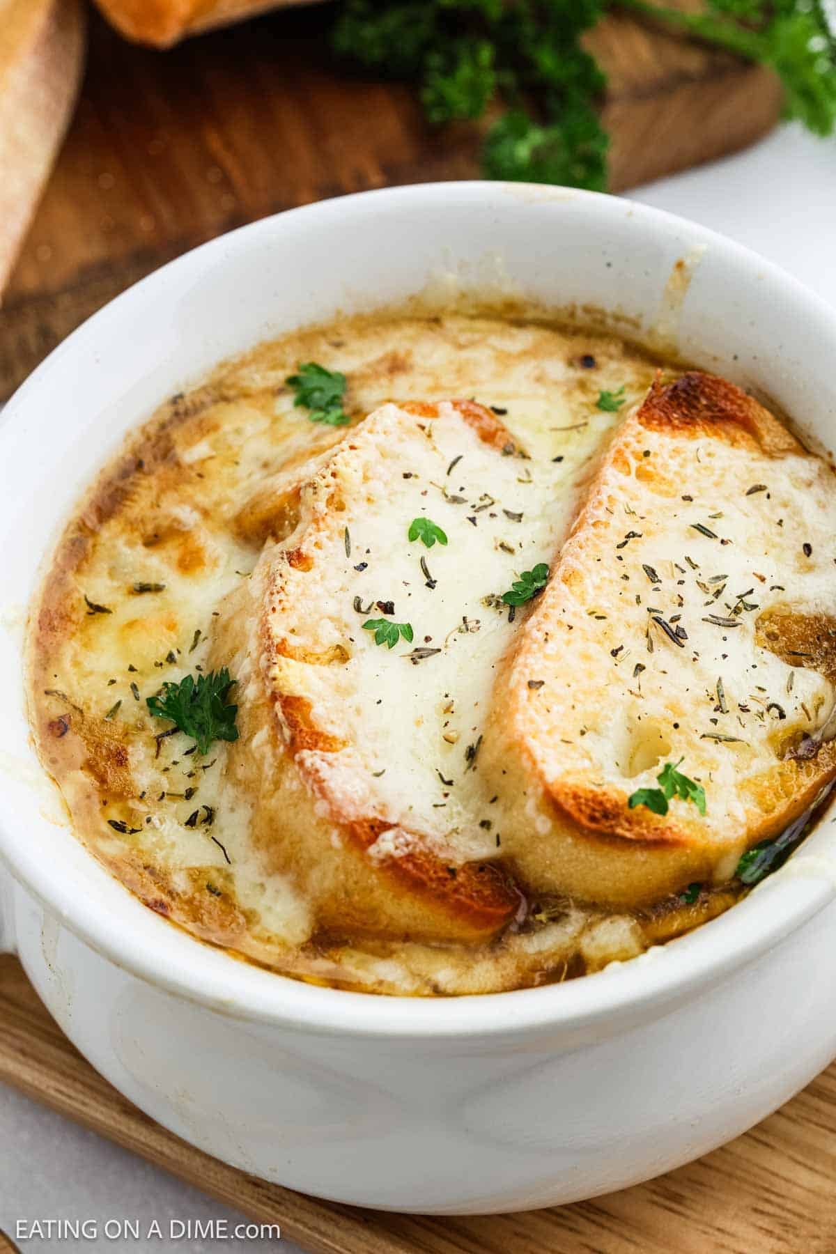 A bowl of classic French Onion Soup topped with two slices of toasted bread and melted cheese, garnished with chopped parsley and herbs.