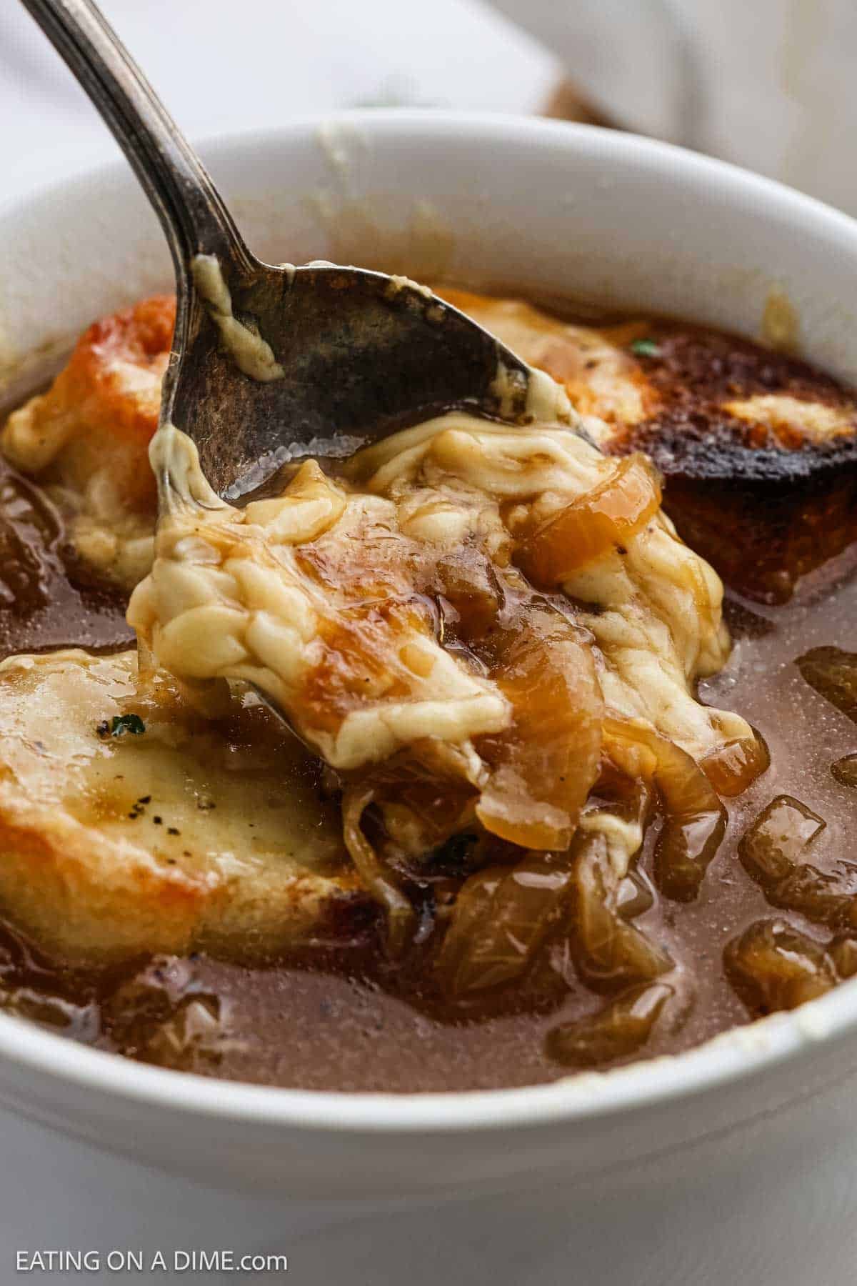 A spoon lifts melted cheese and caramelized onions from a bowl of classic French Onion Soup, revealing toasted bread slices beneath the golden, bubbling cheese.