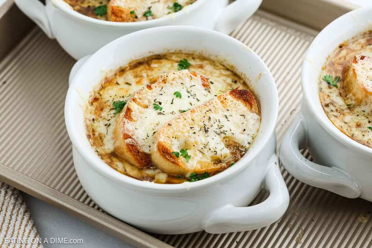 A white bowl of classic French Onion Soup, topped with toasted bread, melted cheese, and sprinkled herbs, sits on a baking tray alongside other bowls of this savory favorite.