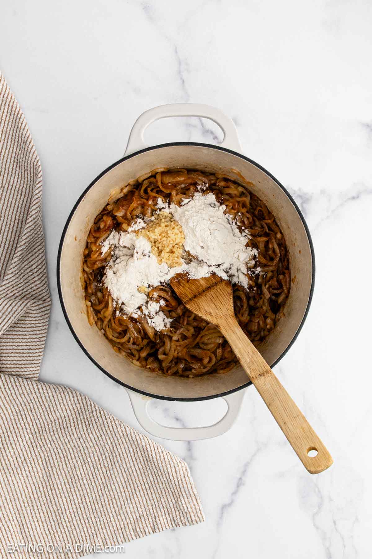 A white pot on a marble surface contains the base for French Onion Soup—sautéed onions, flour, and minced garlic being stirred with a wooden spoon. A beige striped towel rests beside the pot.