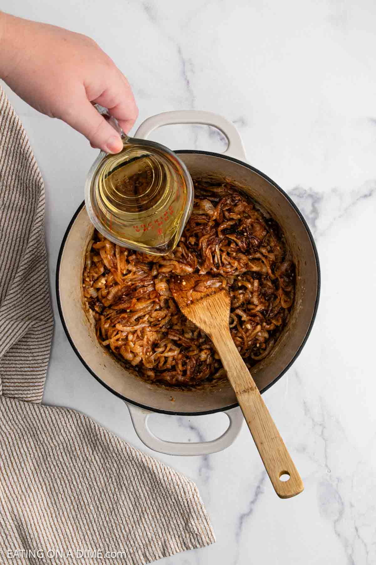 A hand pours liquid from a measuring cup into a white pot filled with caramelized onions for French Onion Soup, using a wooden spoon to stir. The pot sits on a marble countertop next to a striped kitchen towel.