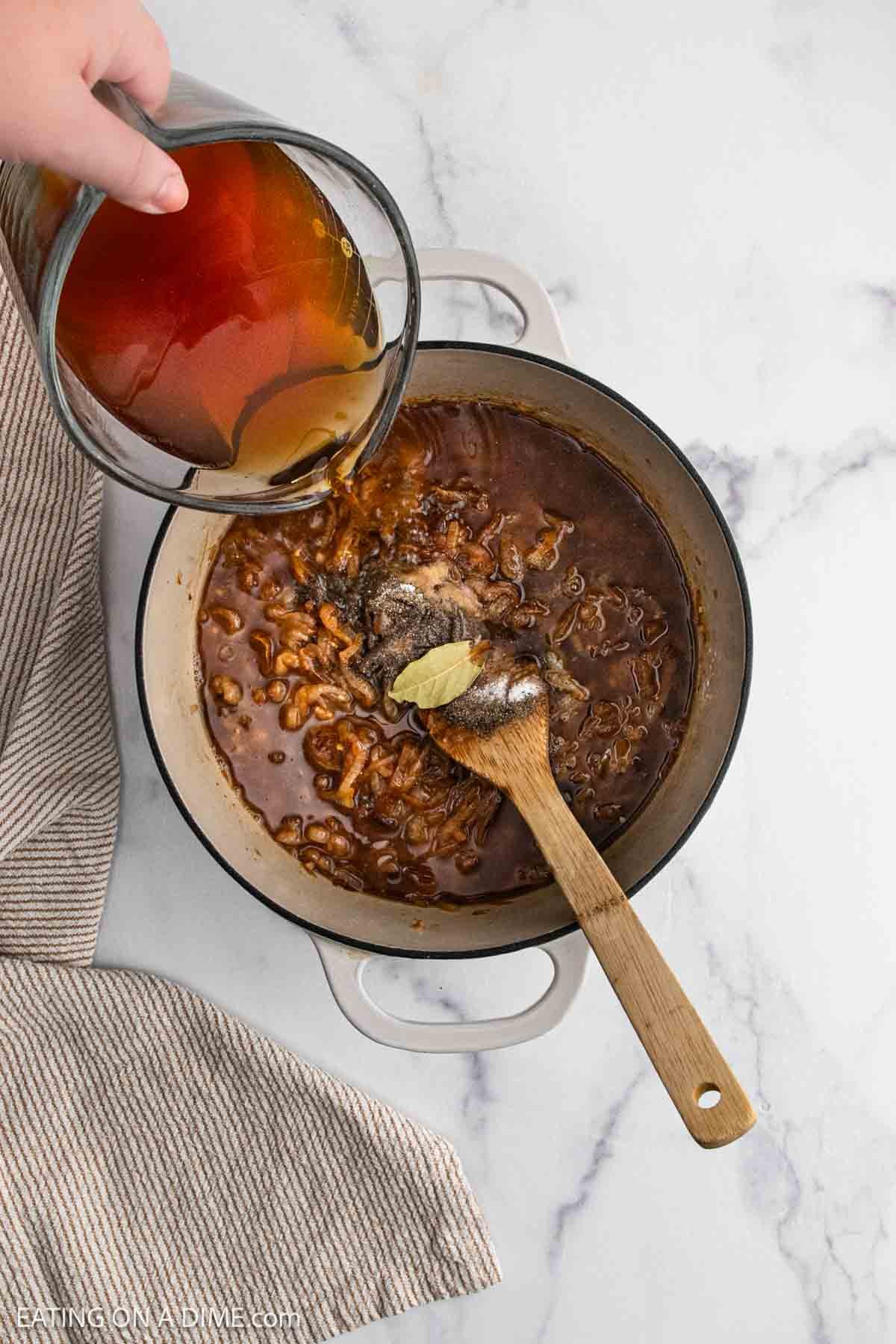 A hand pours broth from a glass measuring cup into a pot of brown ground beef mixture with a bay leaf—evoking the flavors of French Onion Soup—while a wooden spoon rests inside the pot on a marble countertop.