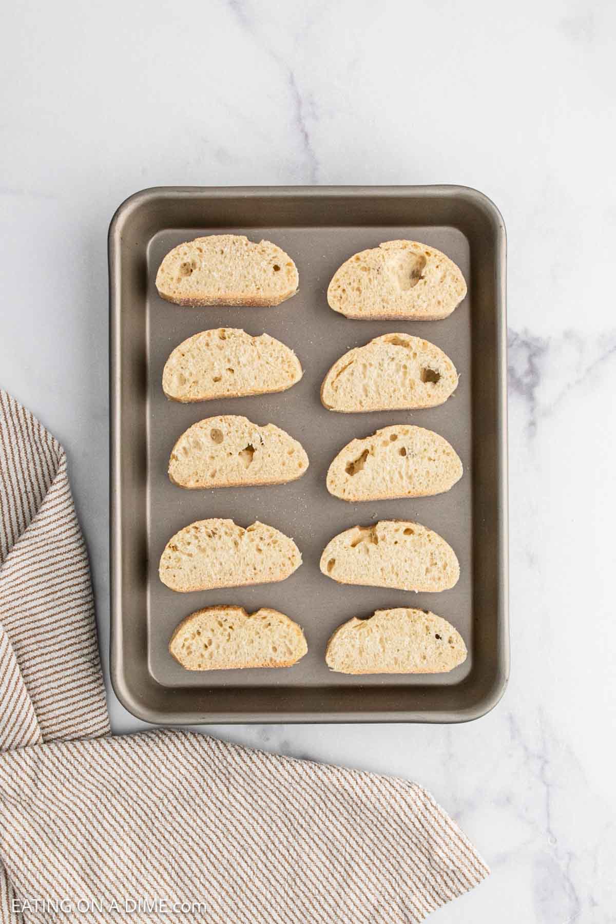 A baking sheet with ten slices of bread, perfect for topping French Onion Soup, arranged in rows on a white marble surface next to a striped kitchen towel.
