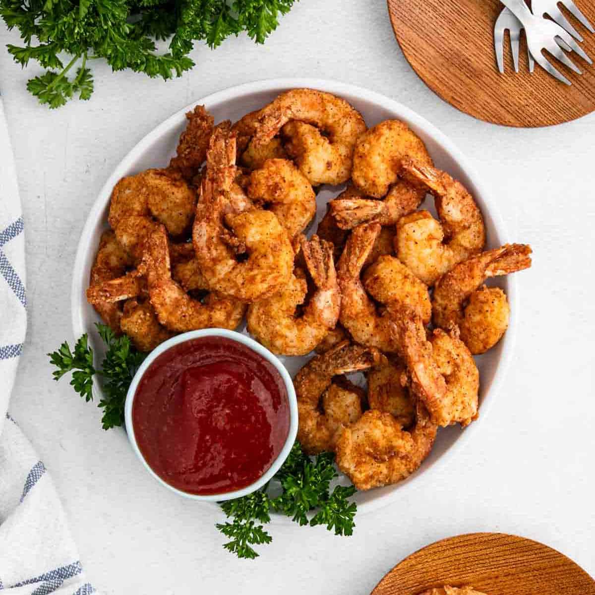 A plate of crispy fried shrimp, a classic in Shrimp Recipes, is served with red dipping sauce and fresh parsley. Wooden plates and utensils complete the inviting presentation on a white surface.