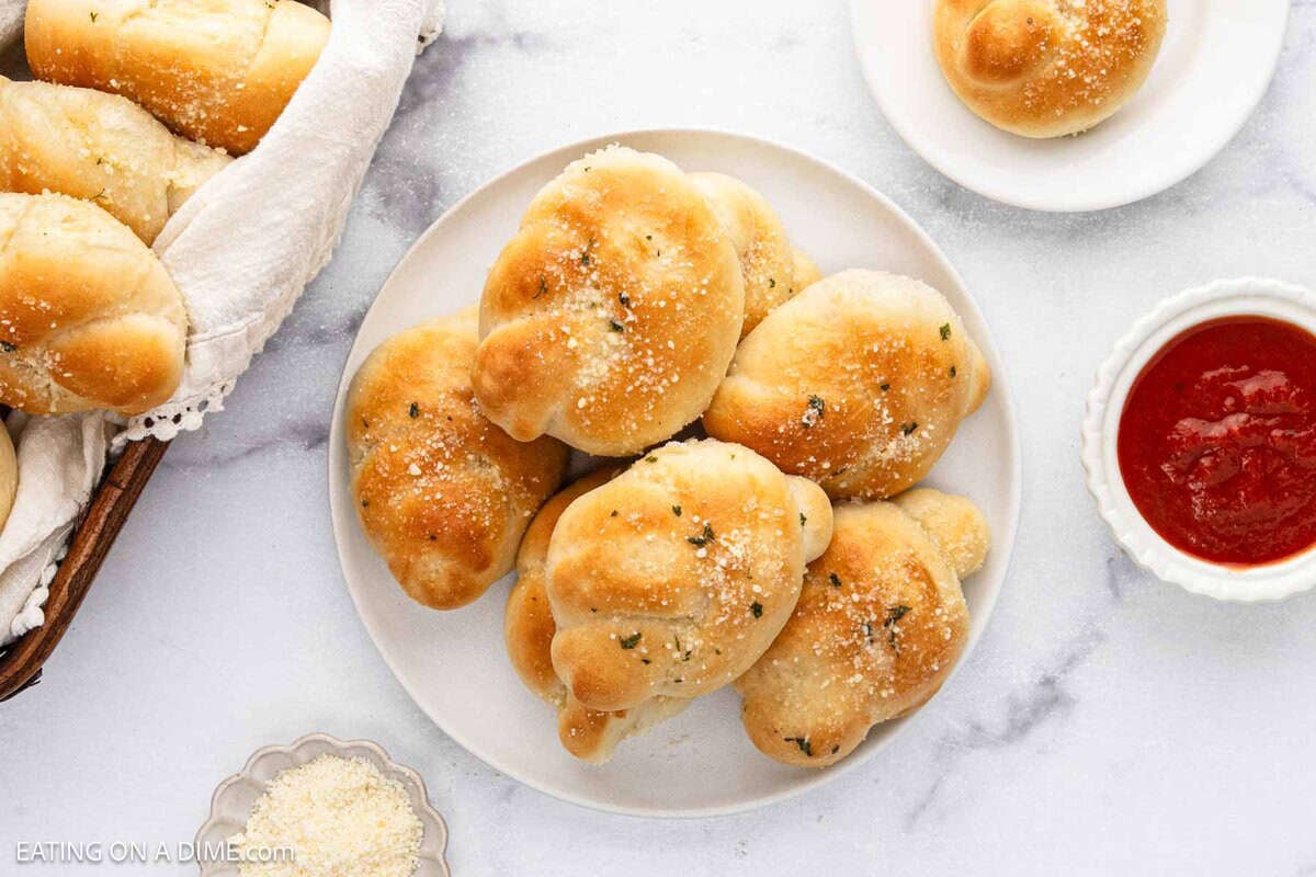 A plate of golden-brown Garlic Knots sprinkled with grated cheese sits on a white surface, accompanied by a bowl of marinara sauce and extra cheese, with more garlic knots in a basket and on a small plate.