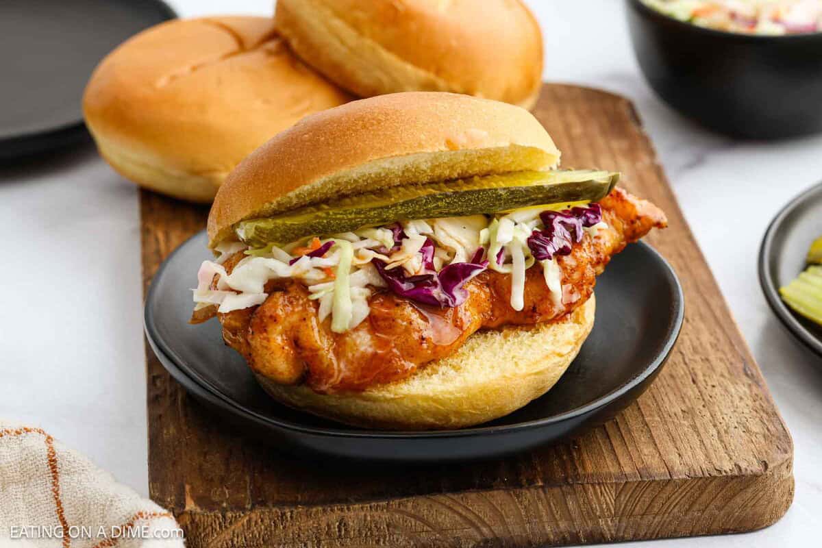 A crispy Hot Honey Chicken Sandwich on a bun, topped with coleslaw and pickle slices, served on a black plate with another bun and a bowl of coleslaw in the background.