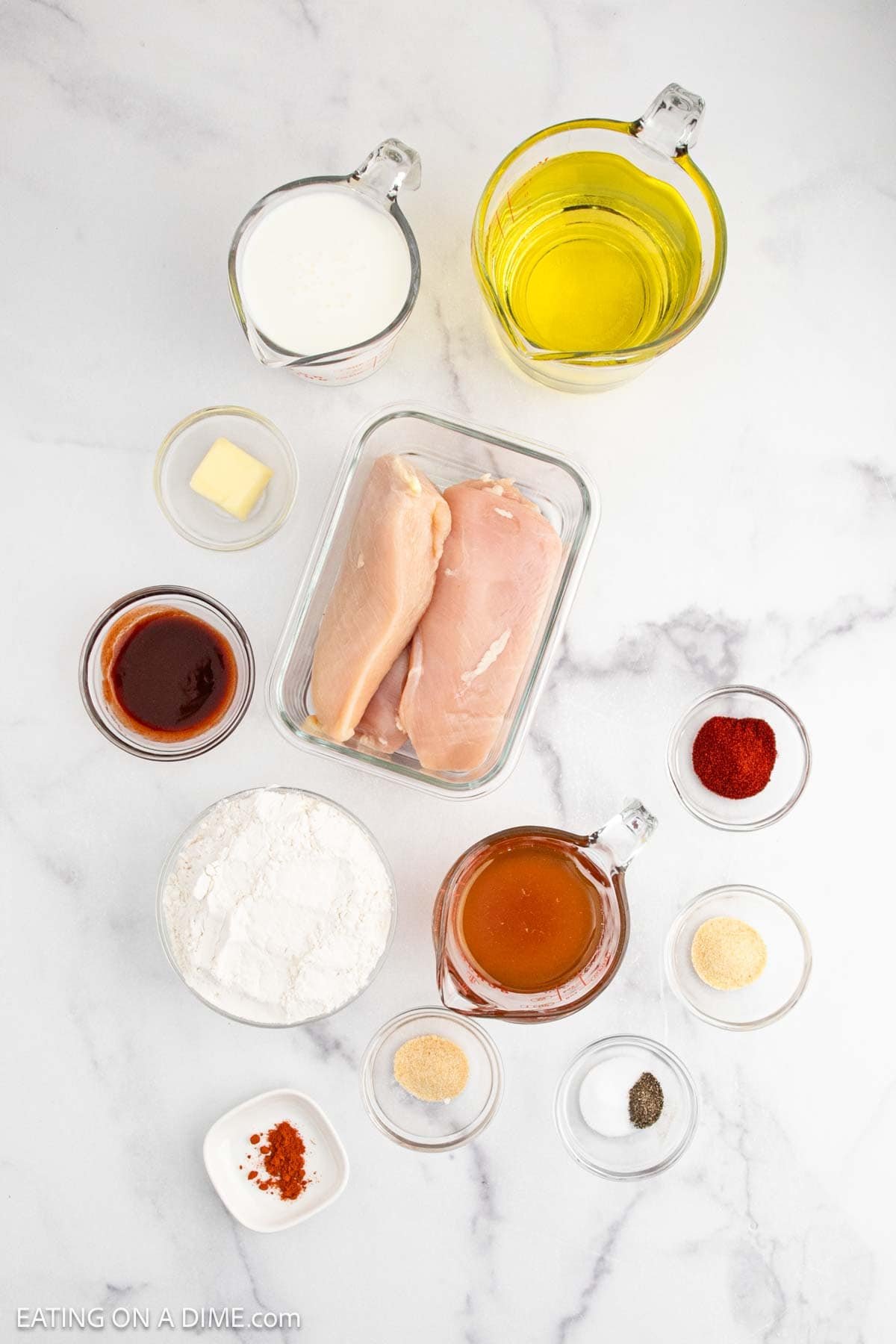 Overhead view of raw chicken breasts, flour, milk, hot sauce, honey, oil, butter, and various spices in small bowls—ingredients for a Hot Honey Chicken Sandwich—arranged on a white marble surface.