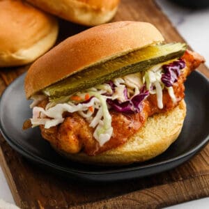 A crispy Hot Honey Chicken Sandwich topped with coleslaw and sliced pickles is served on a black plate, with another sandwich bun visible in the background.