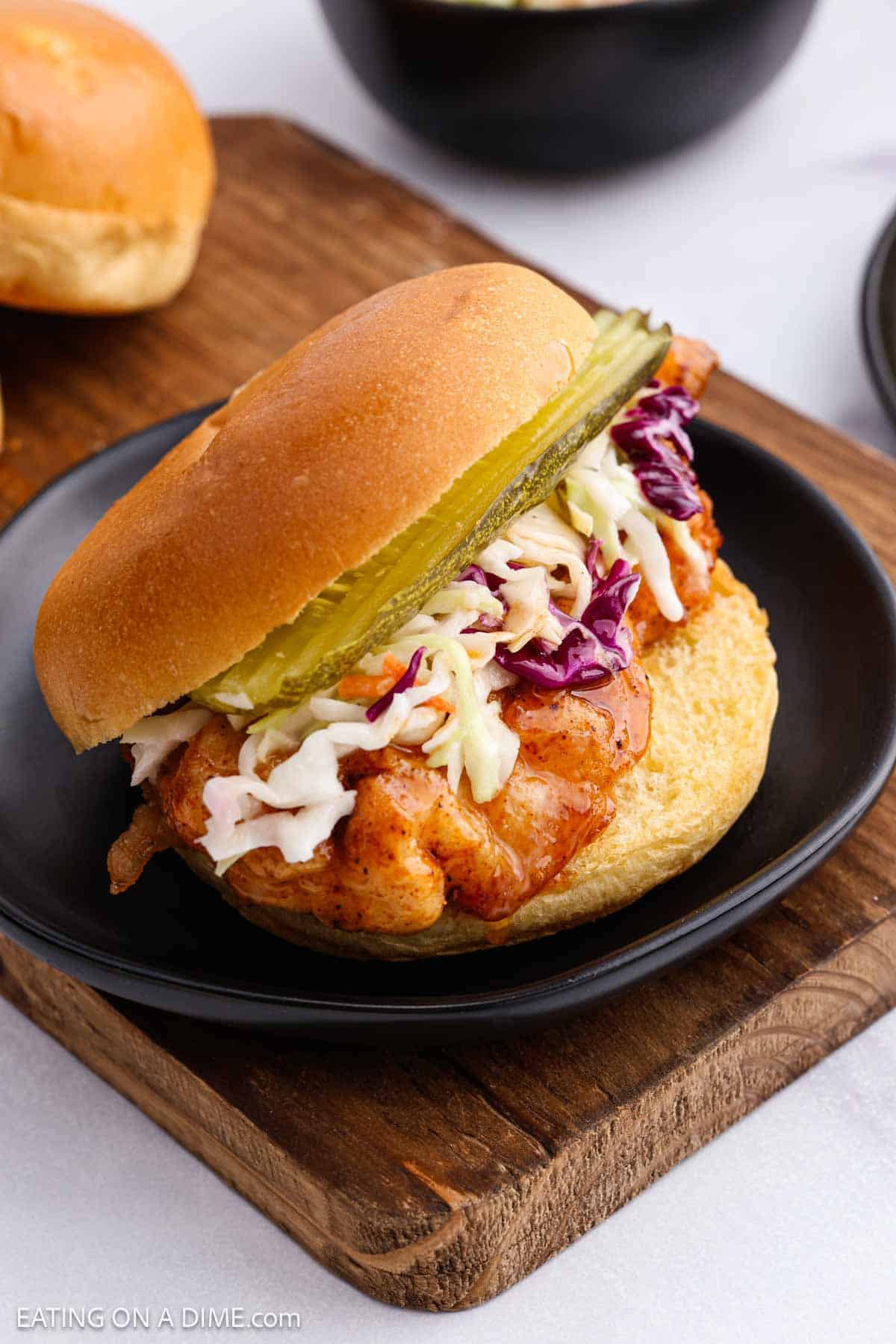 A hot honey chicken sandwich on a black plate, topped with coleslaw and pickle slices, served on a bun. The plate rests on a wooden board with another sandwich visible in the background.