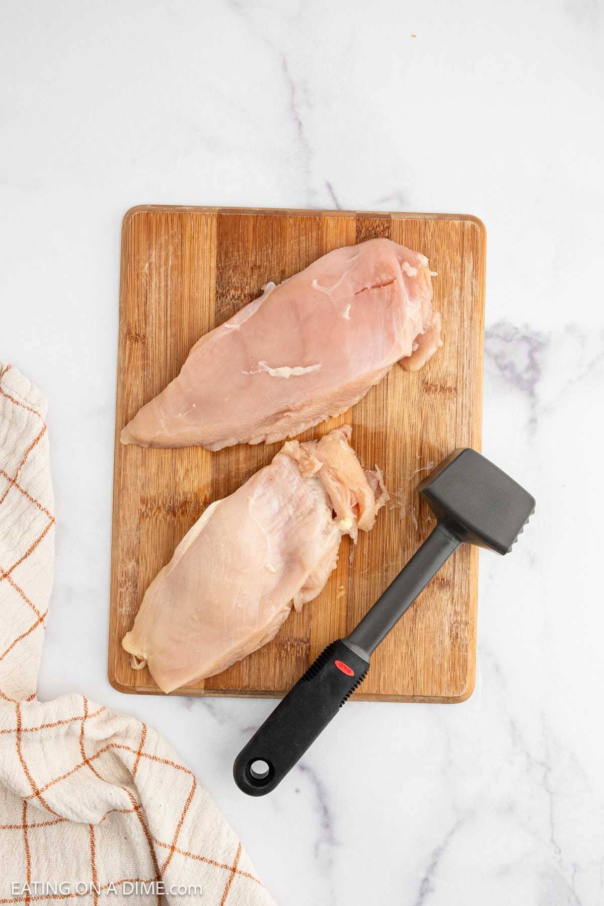 Two raw chicken breasts on a wooden cutting board with a meat tenderizer beside them, ready to be transformed into a delicious Hot Honey Chicken Sandwich, next to a white towel with an orange grid pattern on a white marble surface.