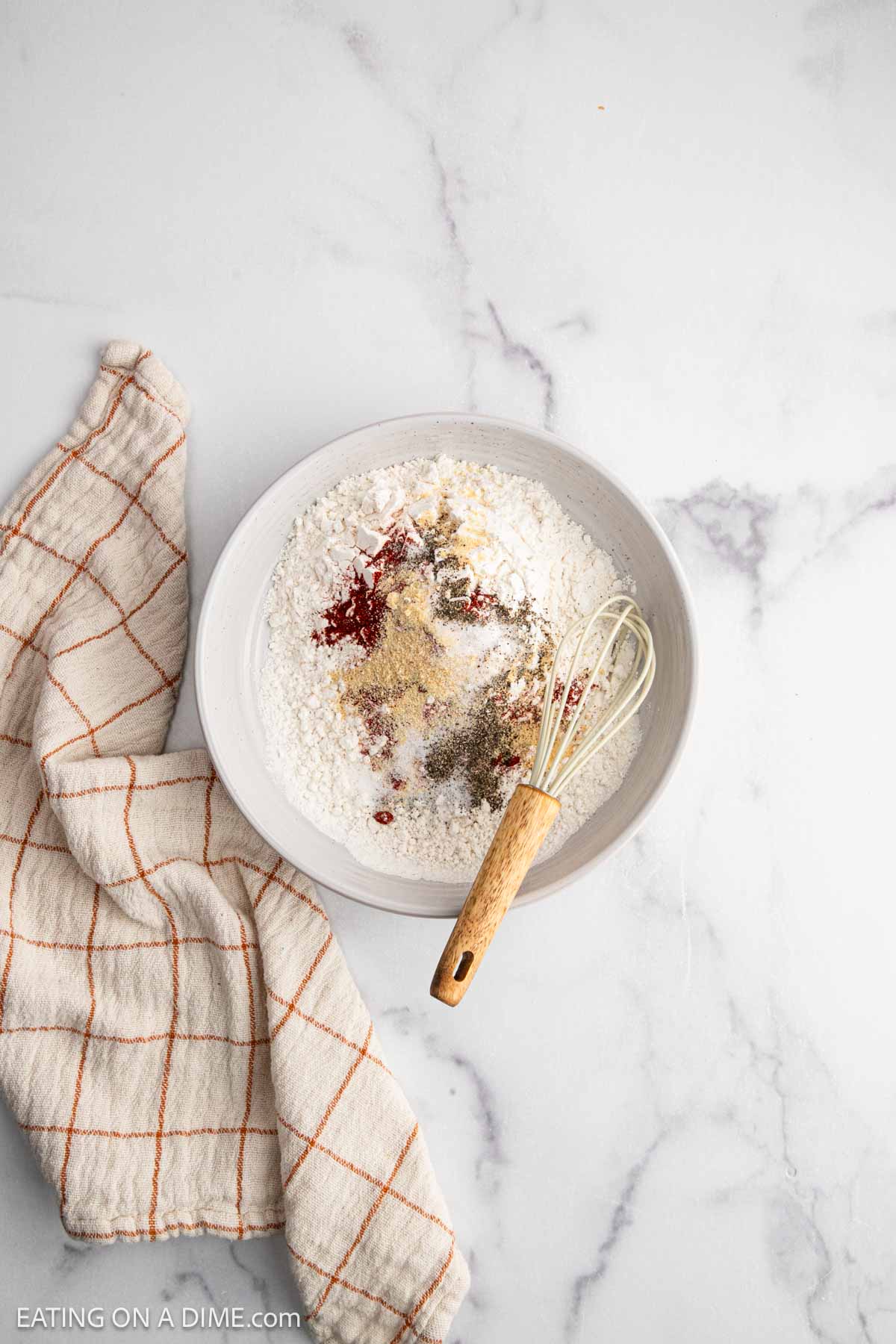 A white bowl filled with flour and assorted spices for Hot Honey Chicken sits on a marble surface next to a beige kitchen towel with orange lines. A whisk with a wooden handle rests inside the bowl, ready for your spicy chicken sandwich prep.