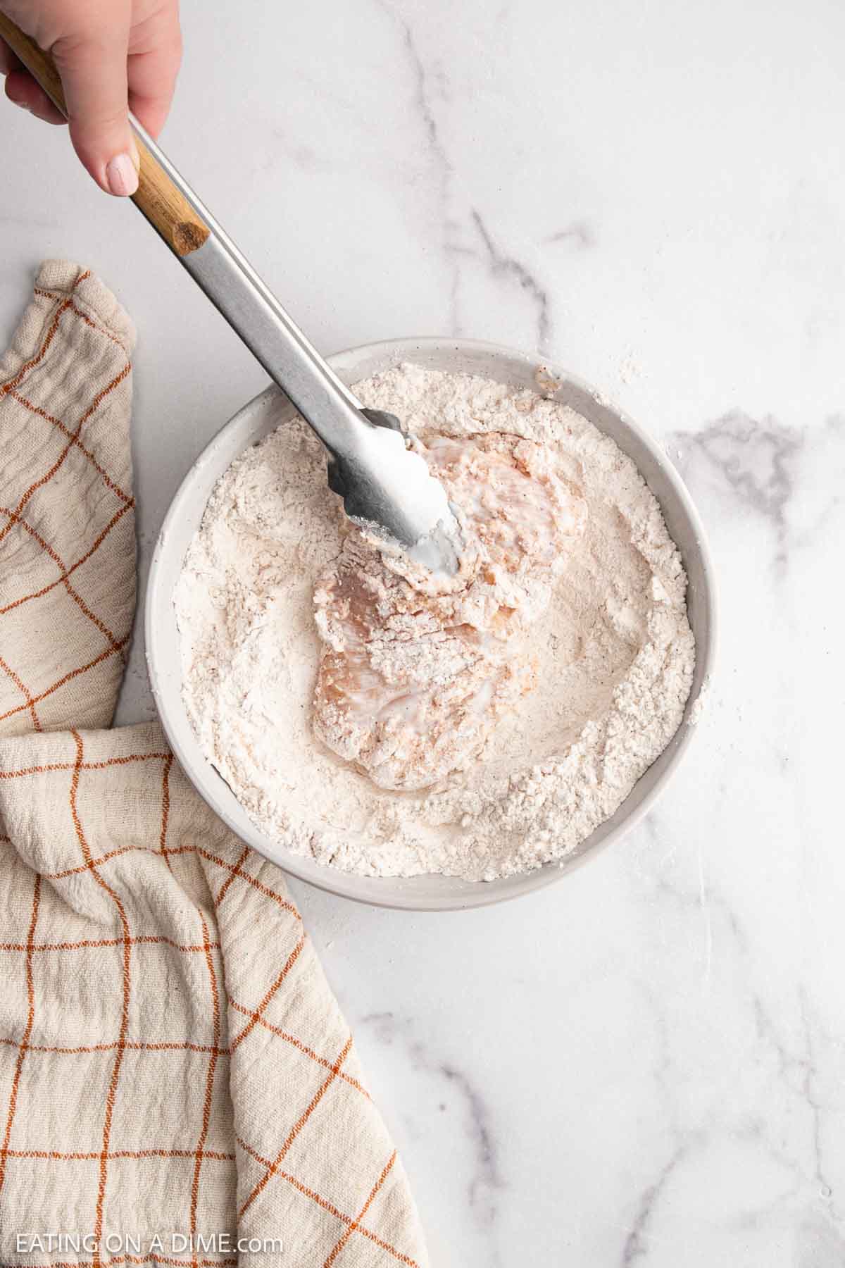 A hand uses metal tongs to coat a raw chicken breast in a white flour mixture inside a bowl, prepping it for a crispy Hot Honey Chicken Sandwich. A beige and orange plaid towel rests nearby on the marble countertop.