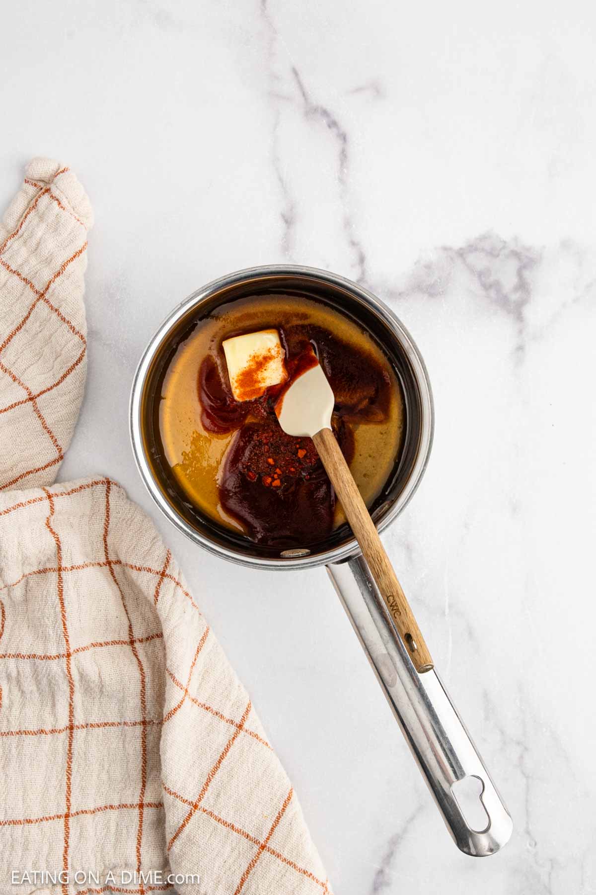 A saucepan with butter, barbecue sauce, hot honey, and spices being mixed with a spatula, next to a beige and orange checkered dish towel on a white marble countertop.
