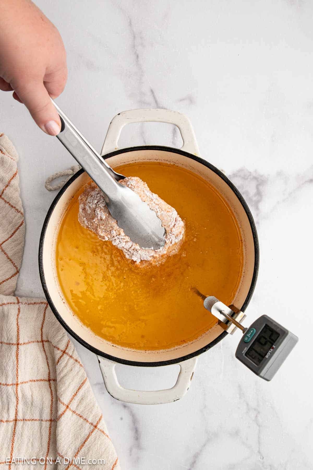 A hand holding metal tongs lowers a piece of breaded Hot Honey Chicken into a pot of hot oil with a cooking thermometer attached. A beige kitchen towel with an orange grid pattern rests nearby on a white surface.