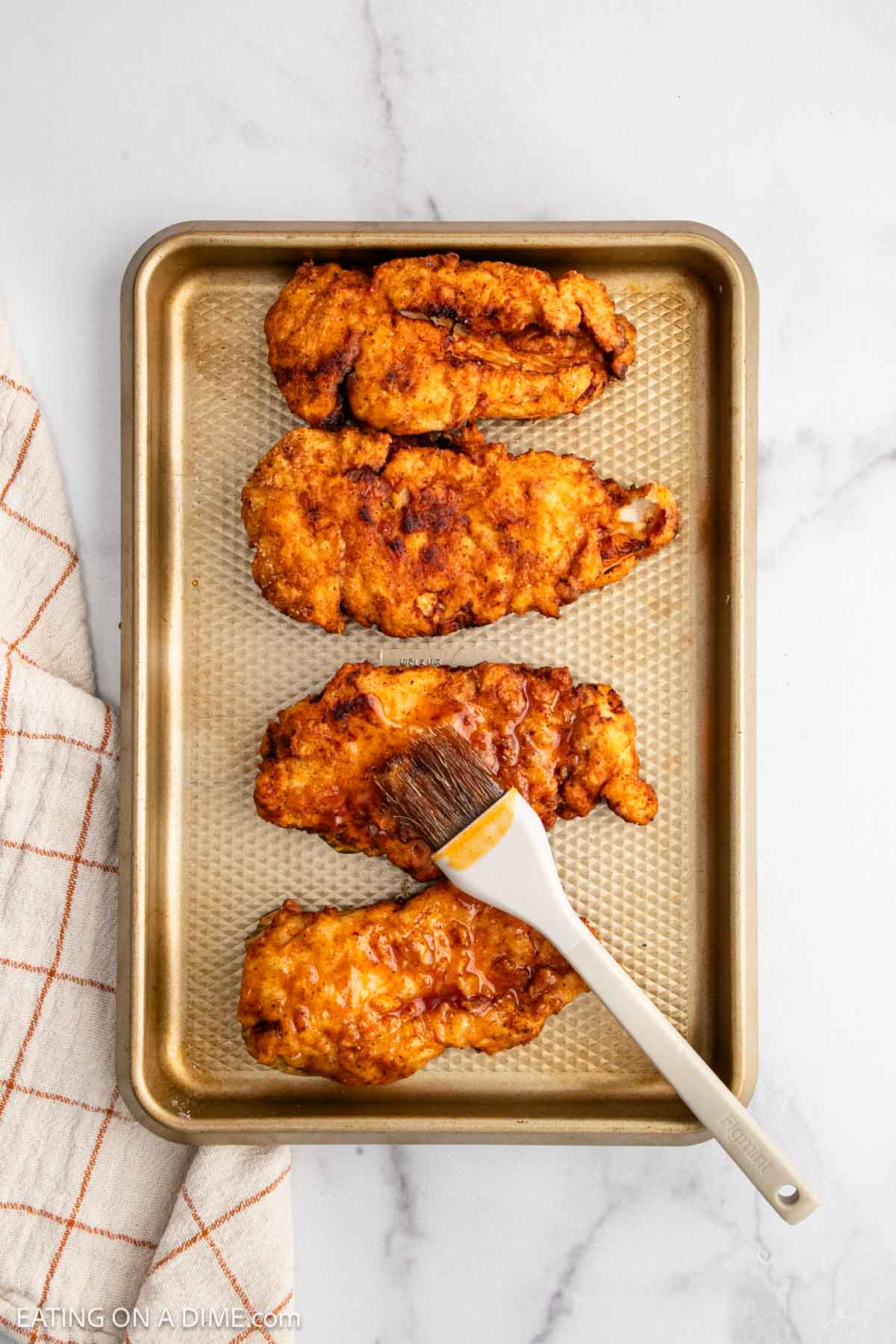 A gold baking sheet holds three pieces of crispy fried chicken, perfect for a Hot Honey Chicken Sandwich. A brush coats one piece with sauce. A beige and orange checked towel sits to the left on a white marble surface.