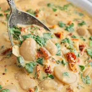 A close-up of creamy Marry Me Butter Beans stew with large white beans, sun-dried tomatoes, wilted greens, and spices, being scooped with a metal spoon.