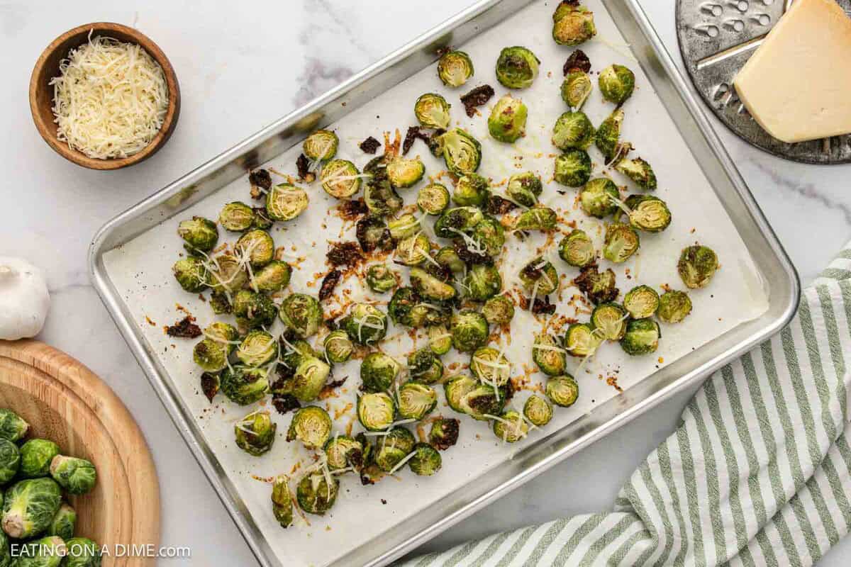 A baking sheet with roasted Parmesan Brussels sprouts topped with shredded cheese, surrounded by a bowl of cheese, a block of Parmesan, a wooden bowl of raw Brussels sprouts, and a green-striped towel.