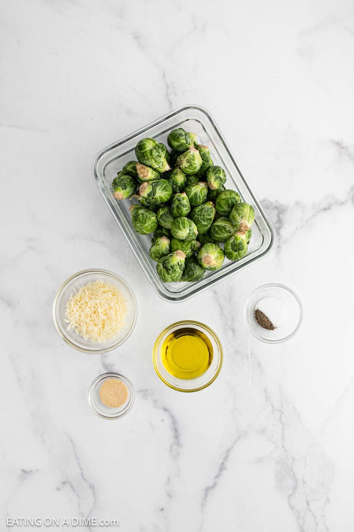 A glass container with fresh Brussel Sprouts, a small bowl of grated Parmesan, a bowl of olive oil, pepper, and garlic powder are arranged on a white marble surface—perfect for making Parmesan Brussel Sprouts.