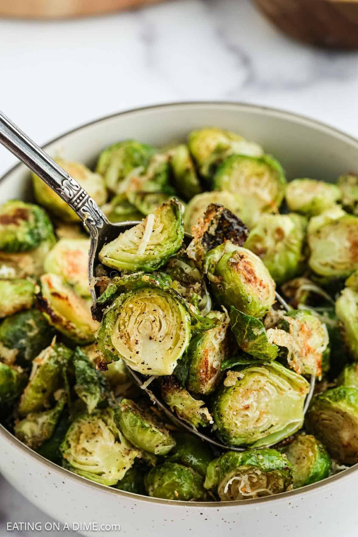 A bowl filled with roasted Parmesan Brussel Sprouts, garnished with grated cheese, with a spoon lifting some of the sprouts.