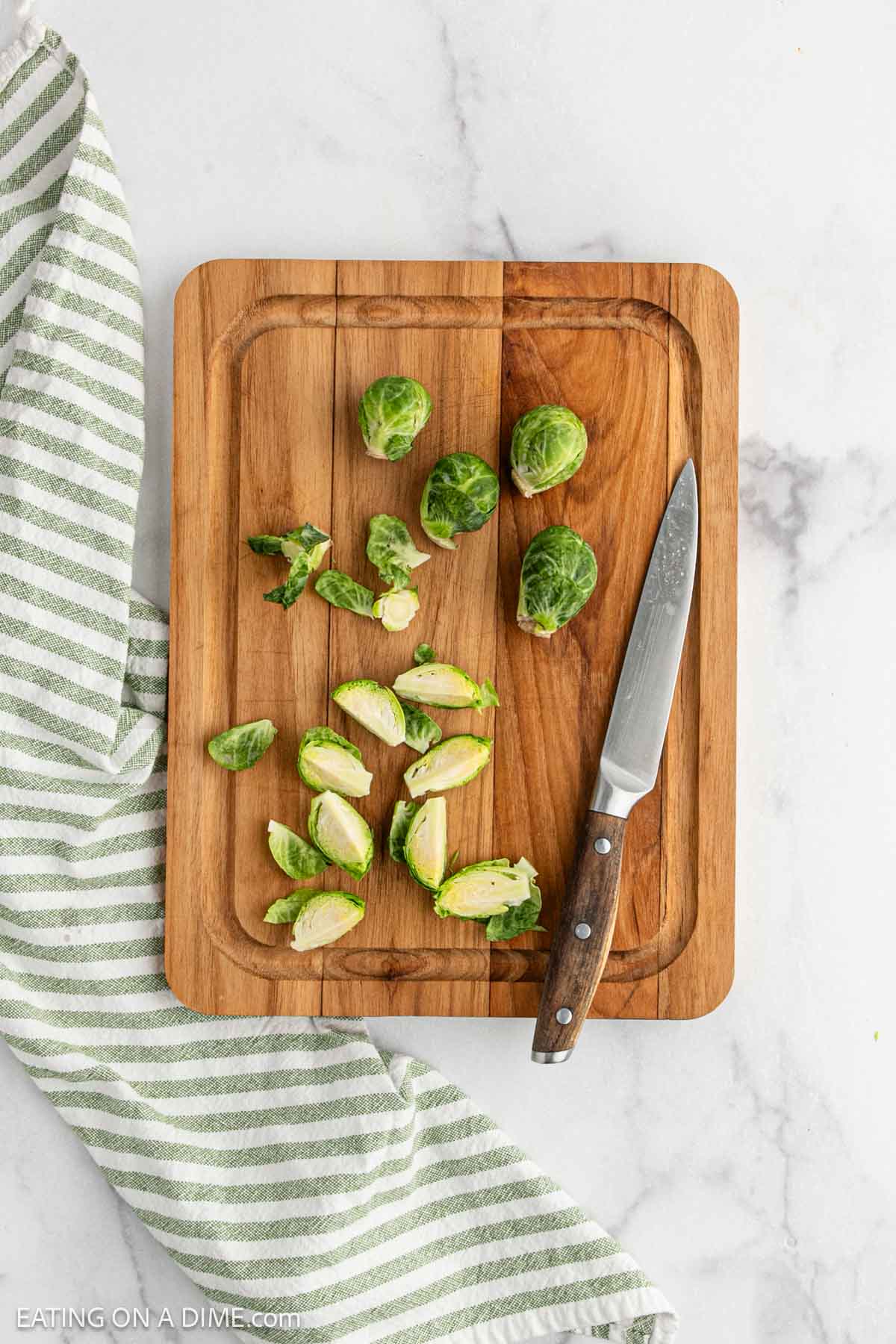 A wooden cutting board with whole and halved Brussels sprouts, ready for making Parmesan Brussel Sprouts, sits beside a paring knife and a green-striped kitchen towel on a marble countertop.