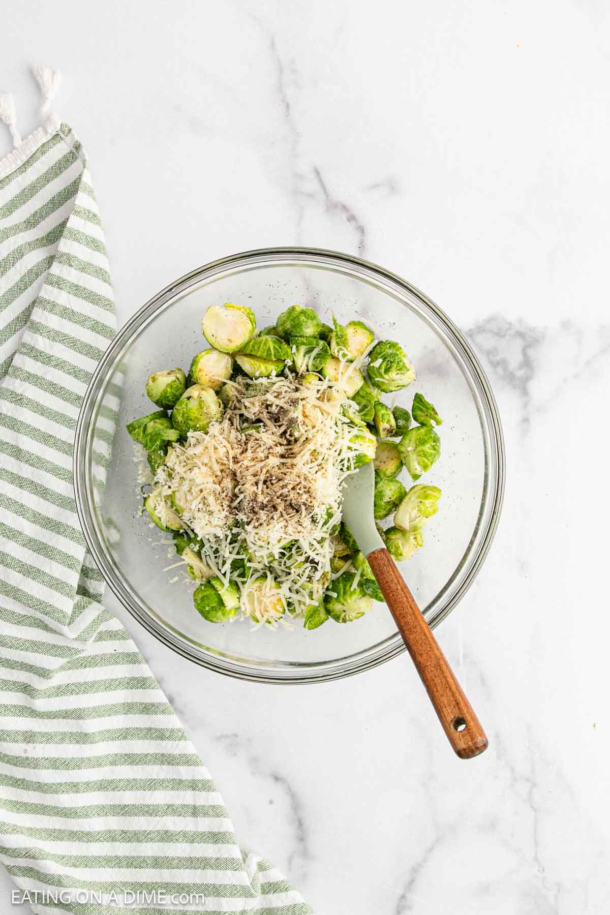 A glass bowl contains halved Brussels sprouts topped with shredded Parmesan and pepper, with a green spatula resting inside. A green-and-white striped towel sits beside this tasty Parmesan Brussel Sprouts dish on a marble surface.