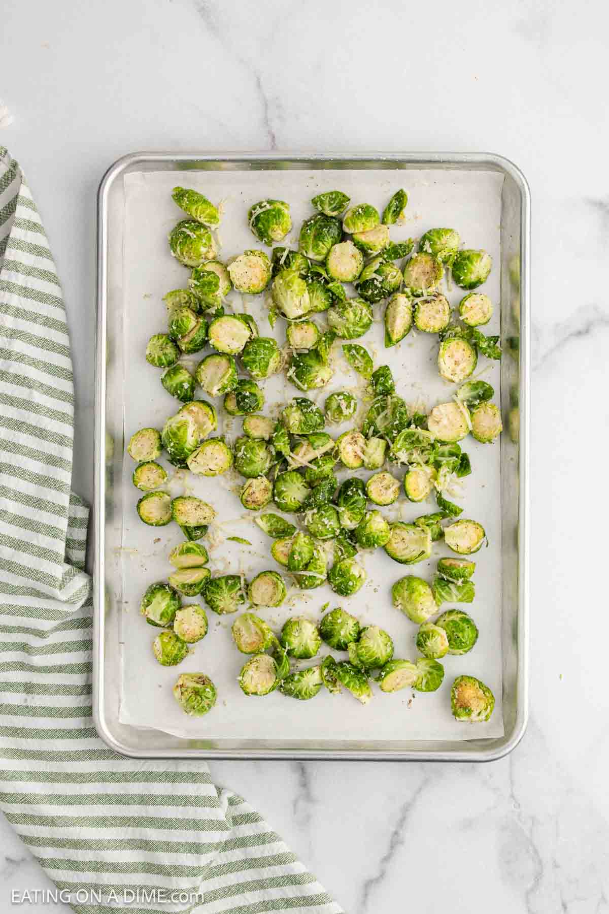 A baking sheet with halved Brussels sprouts sprinkled with seasoning and Parmesan, ready for roasting. A green and white striped cloth is partially visible on the left side of the image.