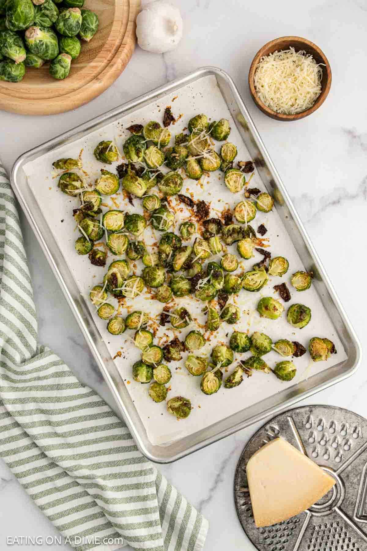 A baking sheet with roasted Parmesan Brussel Sprouts topped with grated cheese sits on a marble surface. Nearby are a bowl of shredded Parmesan, whole Brussels sprouts on a cutting board, garlic, and a striped kitchen towel.