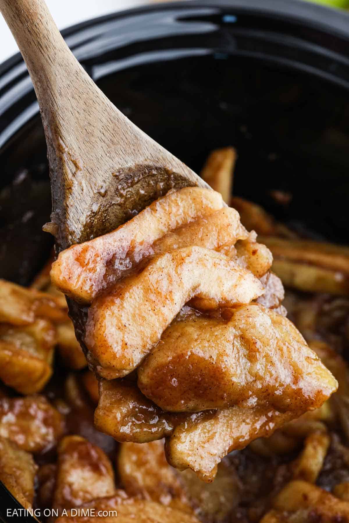 A wooden spoon lifts Slow Cooker Apples—sliced apples coated in a rich, brown cinnamon sauce—from a slow cooker, revealing a warm and gooey apple dessert.