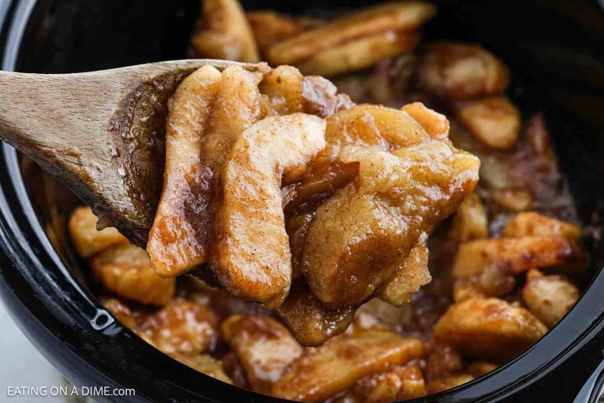 A wooden spoon holds cinnamon-coated apple slices above a pot filled with more cooked apples, suggesting a warm, homemade Slow Cooker Apples dessert or filling.