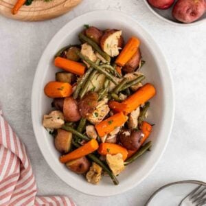 A white oval dish filled with cooked chicken pieces, baby carrots, green beans, and red potatoes, garnished with chopped herbs—a delicious option from Meal Plan 68. A striped towel and a plate with utensils are nearby on a light table.