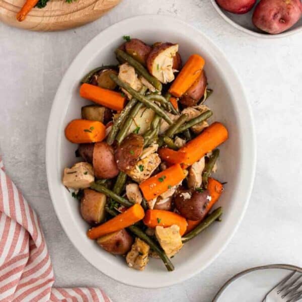 A white oval dish filled with cooked chicken pieces, baby carrots, green beans, and red potatoes, garnished with chopped herbs—a delicious option from Meal Plan 68. A striped towel and a plate with utensils are nearby on a light table.