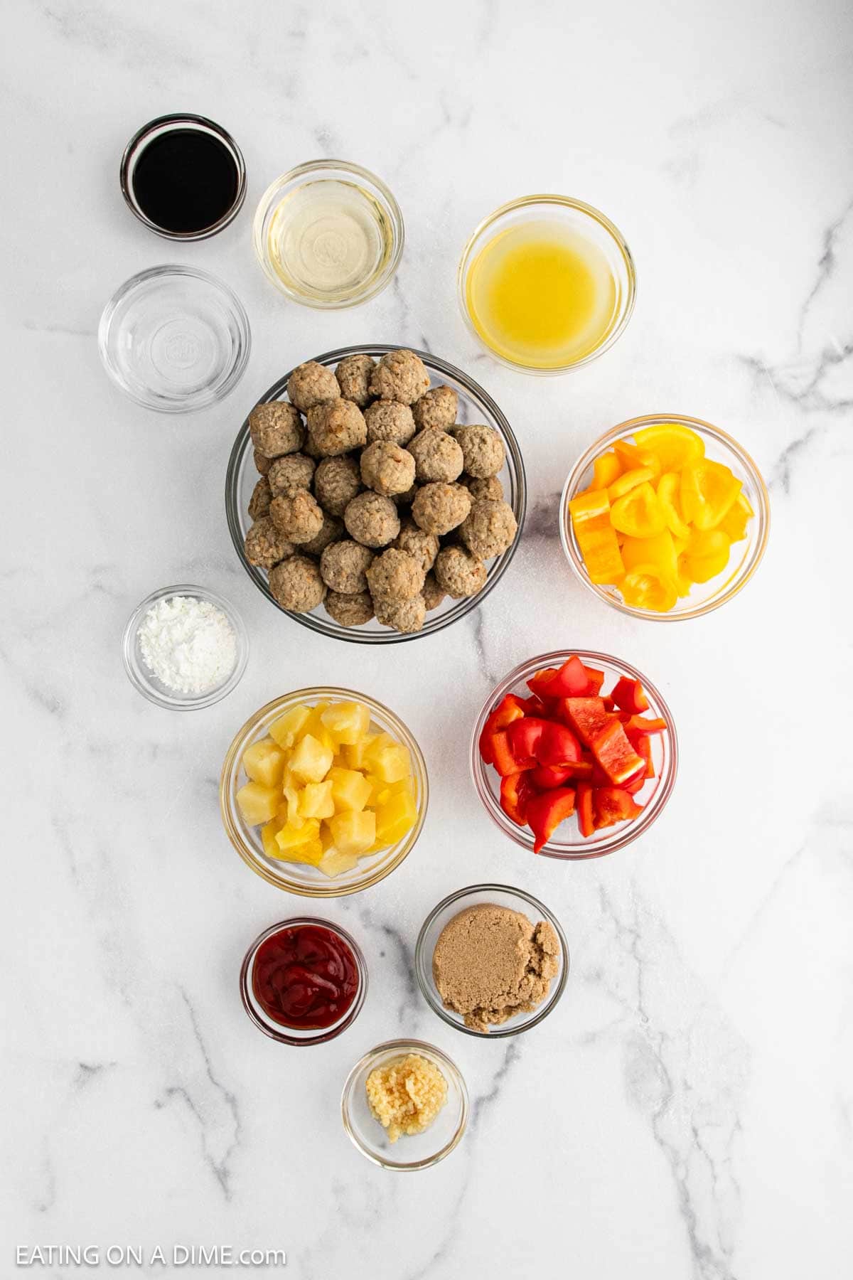 An overhead view of bowls filled with Sweet and Sour Meatballs, pineapple chunks, chopped red and yellow bell peppers, ketchup, brown sugar, minced garlic, cornstarch, and various sauces on a white marble surface.