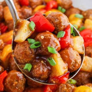 A close-up of sweet and sour meatballs with pineapple chunks, red and yellow bell peppers, and green onion slices, all coated in a glossy sweet and sour sauce. More meatballs and vegetables are visible in the background—a mouthwatering meatballs recipe.