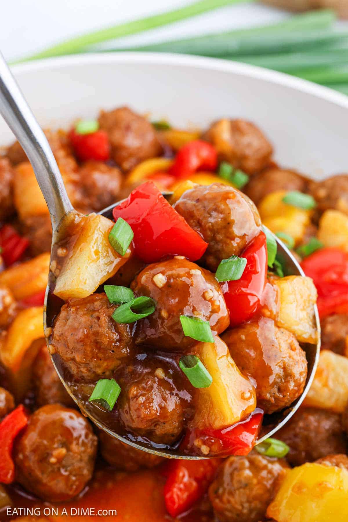 A close-up of a spoon lifting Sweet and Sour Meatballs with chunks of red and yellow bell peppers, pineapple, and glossy sauce, garnished with sliced green onions.