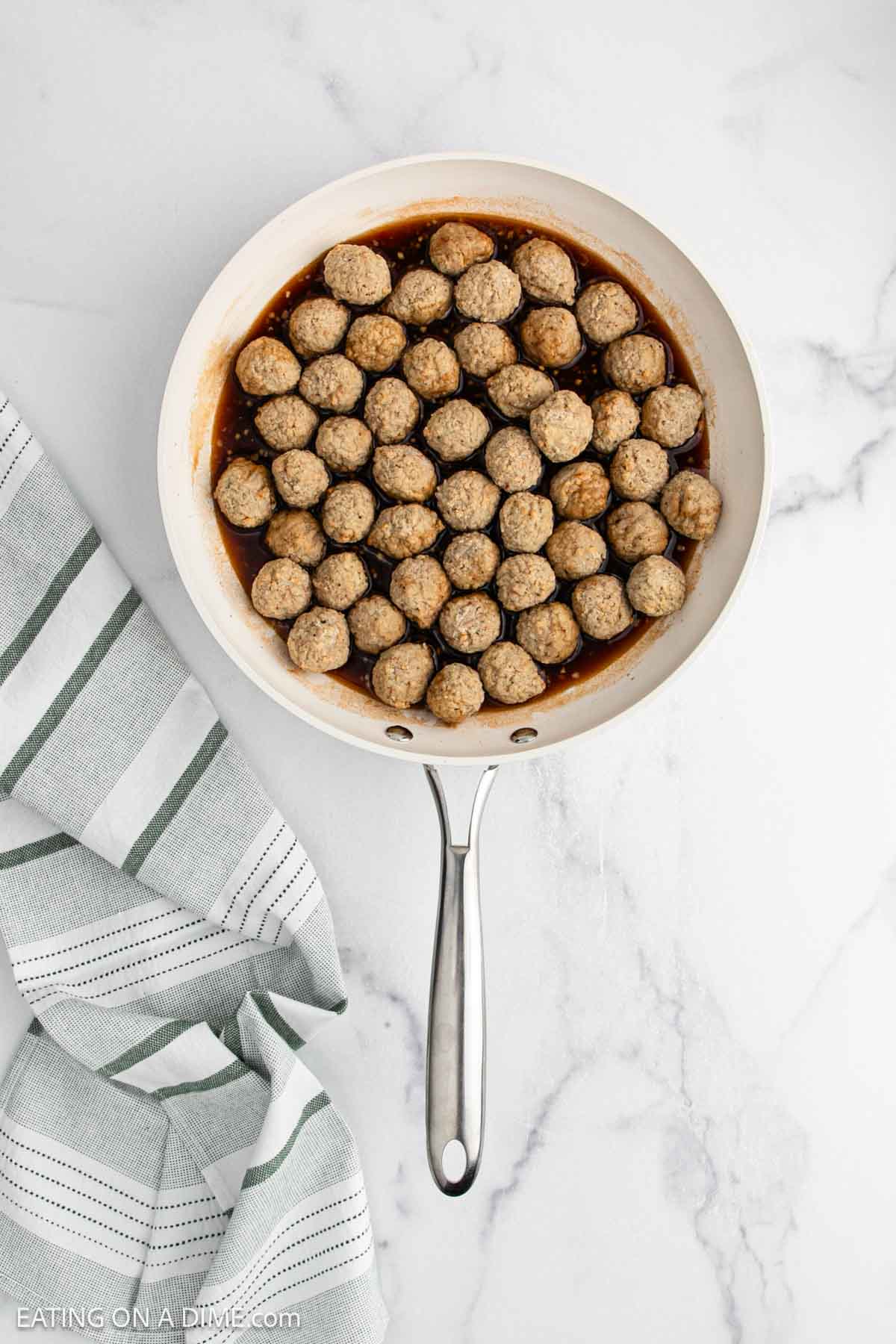 A white skillet filled with evenly arranged sweet and sour meatballs in sauce sits on a white marble surface next to a green and white striped kitchen towel.