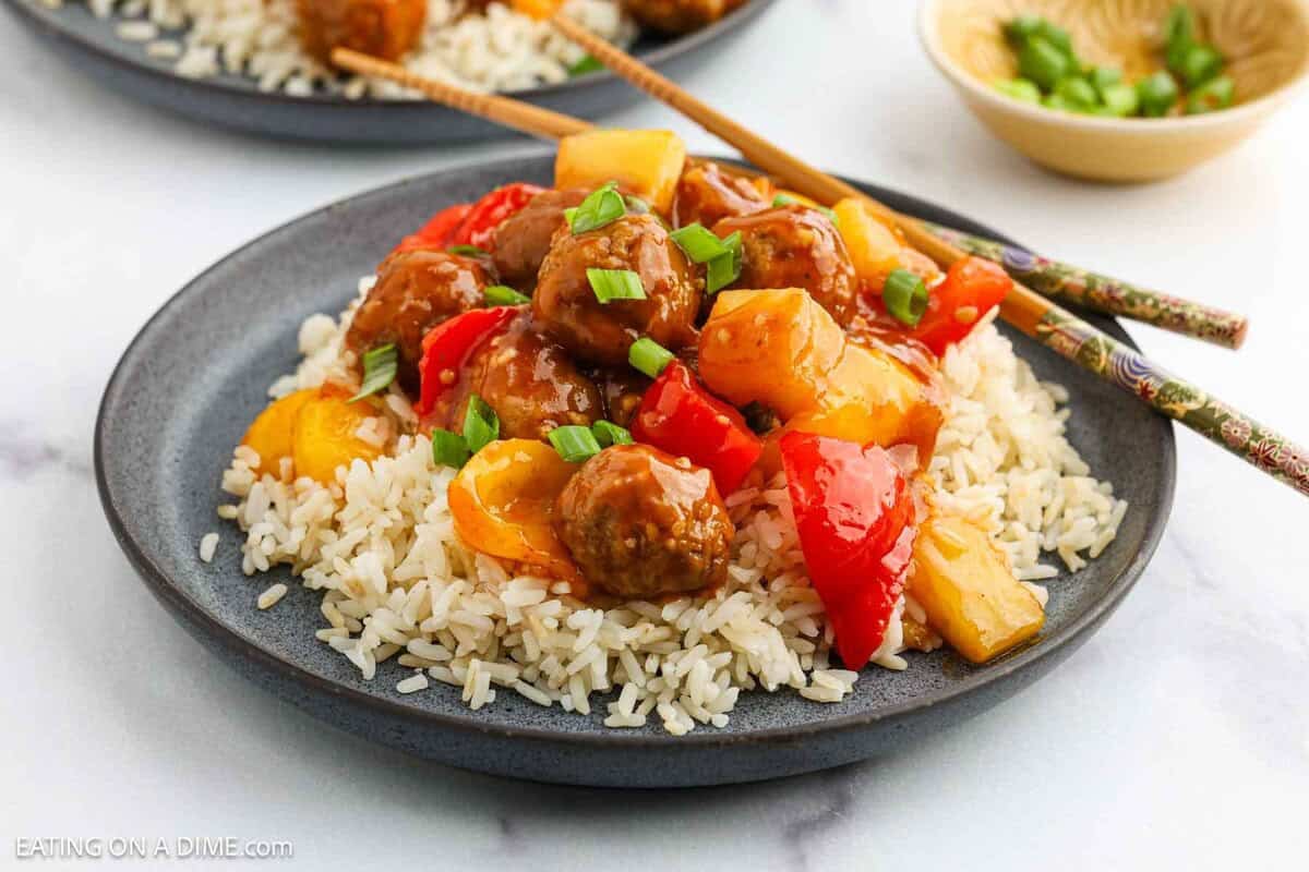 A plate of white rice topped with Sweet and Sour Meatballs, colorful bell peppers, pineapple chunks, and chopped green onions. Chopsticks rest on the side of this vibrant Sweet and Sour Meatballs dish.