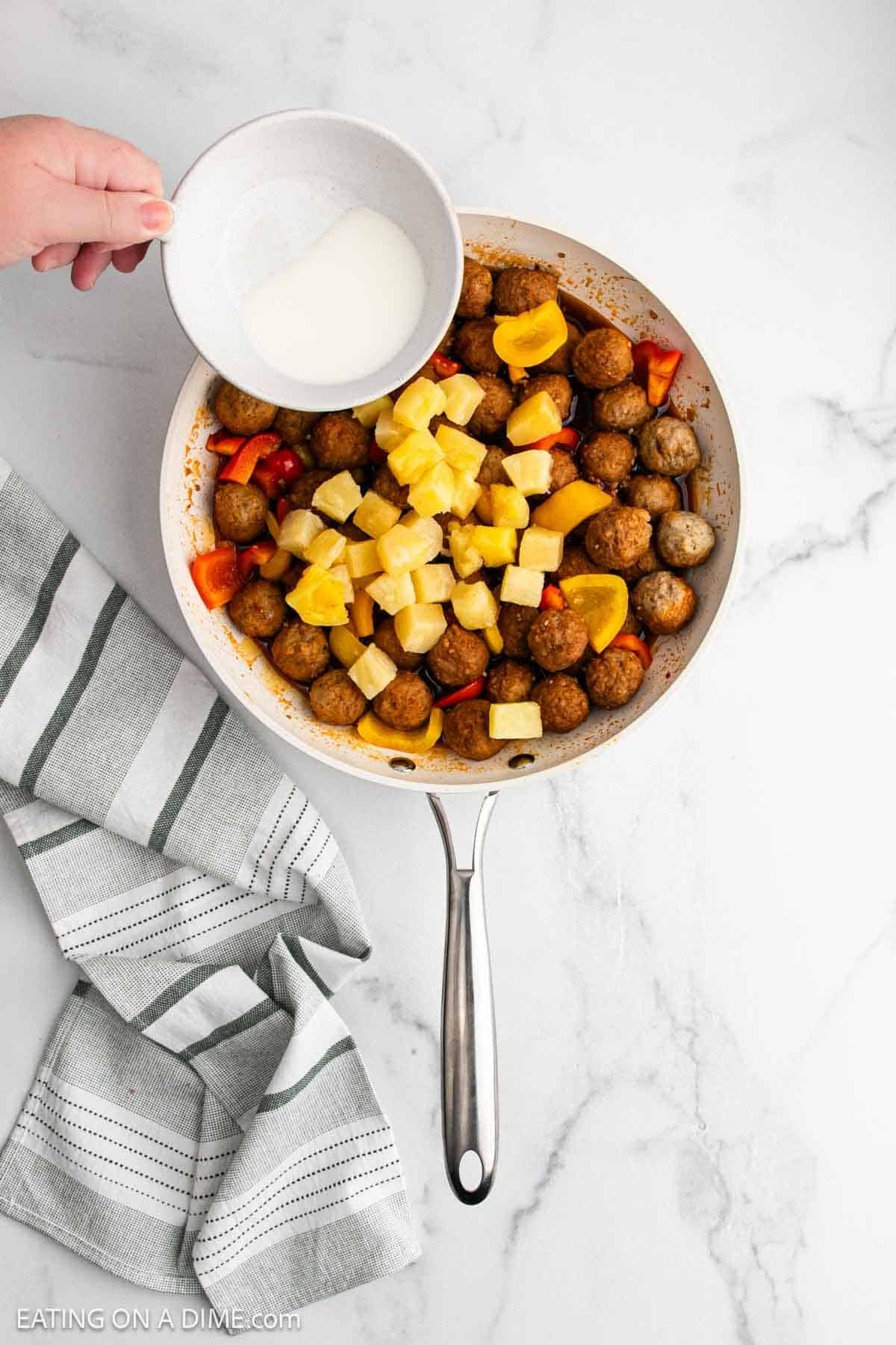 A hand pours a small bowl of milk into a skillet filled with Sweet and Sour Meatballs, pineapple chunks, and red bell peppers. A striped kitchen towel rests beside the skillet on a white marble surface.