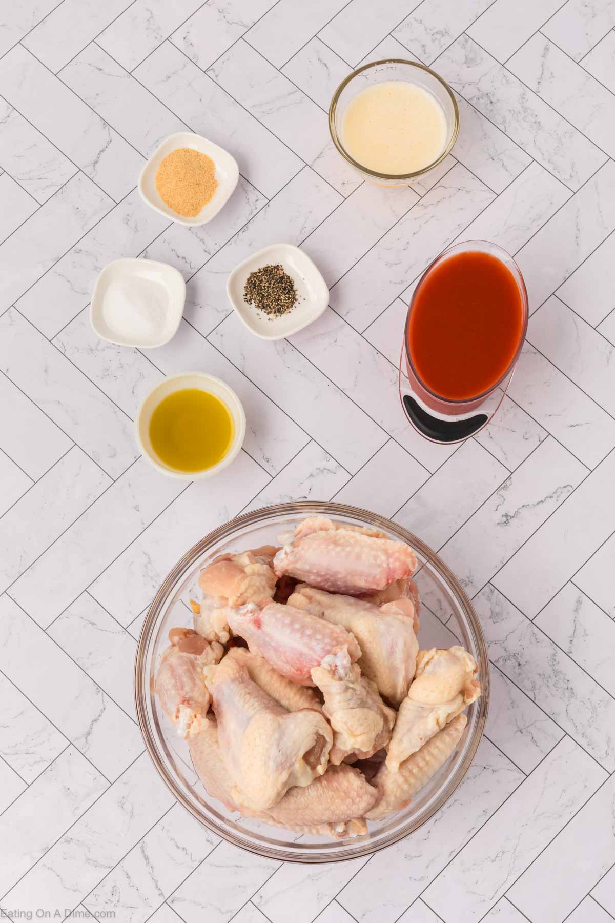 A glass bowl of raw chicken wings, perfect for making baked chicken wings, sits surrounded by small bowls of hot sauce, olive oil, black pepper, salt, garlic powder, and a light-colored liquid on a white herringbone-patterned surface.
