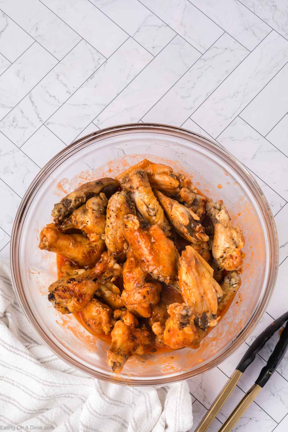 A glass bowl filled with saucy, baked chicken wings sits on a white tiled surface next to a cloth napkin and a pair of metal tongs.